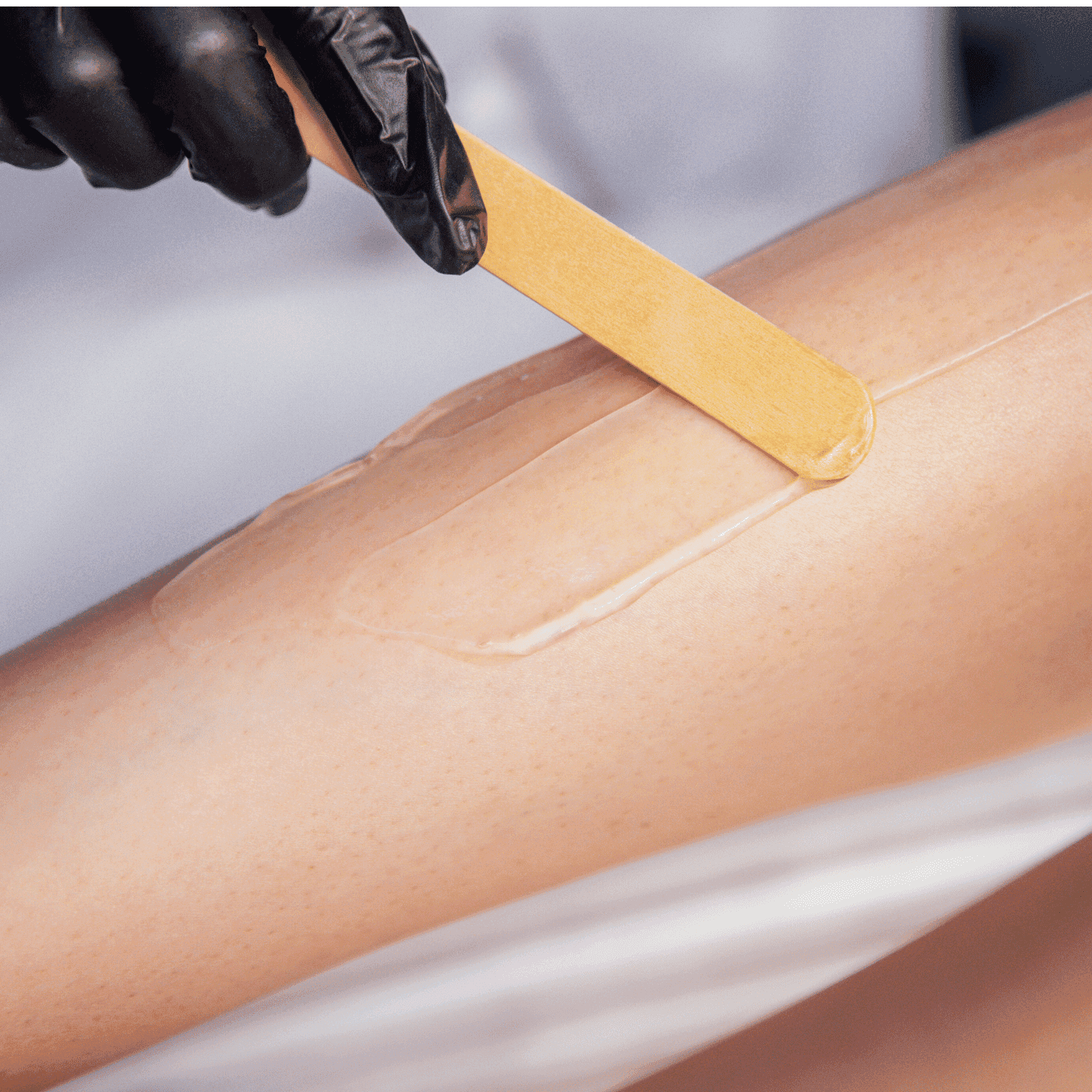 Leg being waxed by person with gloved hand, applying wax strip with wooden spatula.